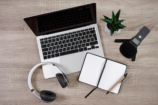 journalism, podcast and blogging concept - top view of workplace with laptop, microphone, headphones, open notepad and pen over wooden table background