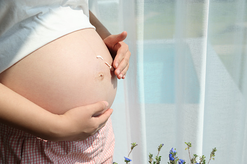 Pregnant woman applying stretch mark cream on her belly by the window at home