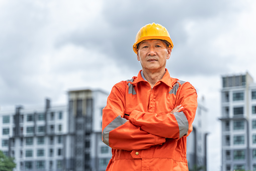 Serious and determined middle age Asian engineer in orange jumpsuit with hard hat crossing his arms while standing proudly in front of his multi-stories buildings with cloudy sky in the background
