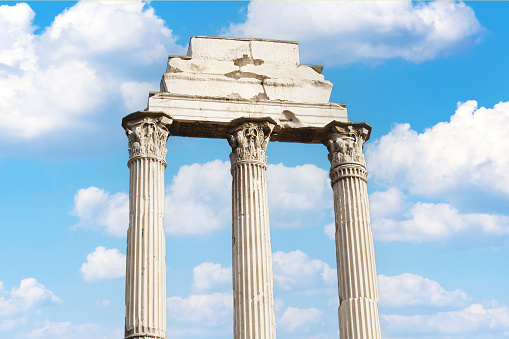 Three Roman temple pillar ruins in Rome, Italy against a cloudy blue sky.
