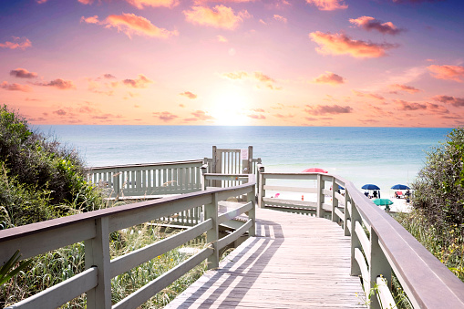 A low angle view down a wooden public boardwalk leading to the beach and ocean in Rosemary Beach, Florida during a vibrant sunset.