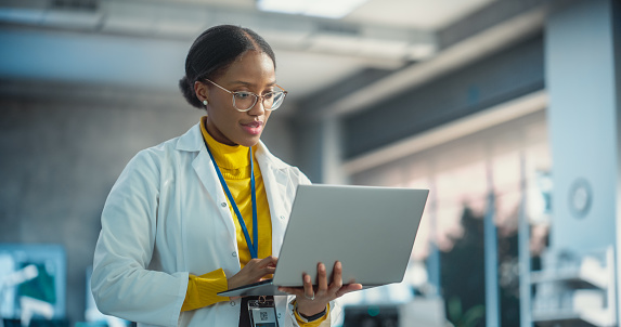 Portrait of Young Black Female Specialist in a Lab Coat Using Laptop. Professional and Successful Woman Working as an Engineer, Thinking and Developing High Technology Projects In Startup