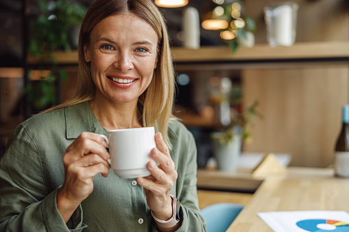 Attractive female manager smiling and looking at camera while drinking coffee in cozy cafe