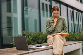 Pensive woman entrepreneur working on laptop outside on modern building background and making notes