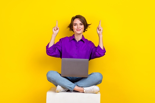 Full size photo of pretty positive girl sit podium direct fingers up empty space use netbook isolated on yellow color background.