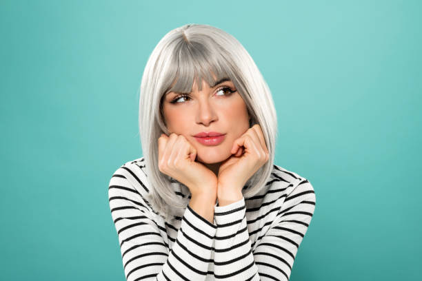 Dreamy female model with hands at chin looking away Pensive young woman in gray wig and striped black and white shirt looking away thoughtfully while sitting with hands at chin against turquoise background gray hair young woman stock pictures, royalty-free photos & images