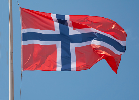 Norway the Norwegian national flag waving in the wind on a flagpole with blue sky background.