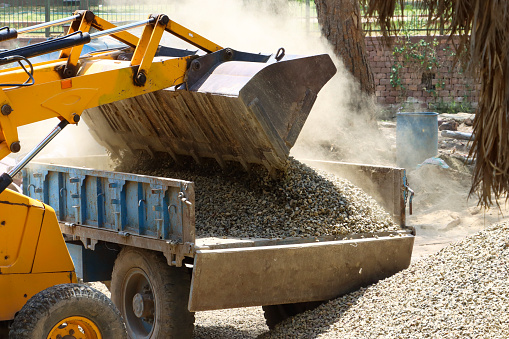 Stock photo showing close-up view of heap of gravel on construction site being transported by the toothed bucket of a wheeled excavator into the back of a flat bed lorry.
