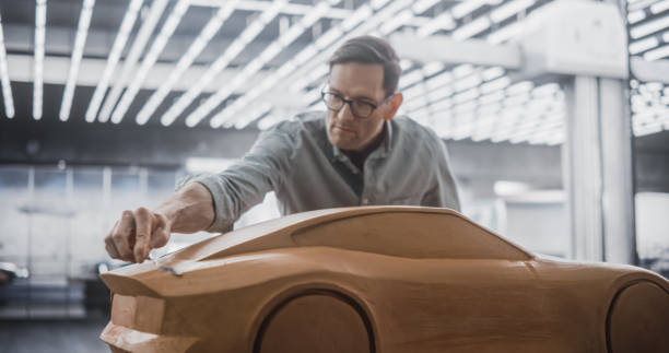 istock Car Modeler Enjoying Creative Work Process on a Concept Car Made Out of Plasticine Modeling Clay. Handsome Designer Using Spatula to Smooth the Prototype Model of a Modern Electric Vehicle 1516861723