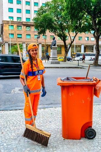 Rio de Janeiro, Brazil - June 15, 2023: A female street sweeper holding a broom and smiling to the camera. She is in orange high-visibility clothing.