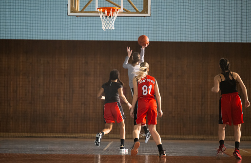 Image from a low angle viewpoint of a basketball player going to shoot a ball in a hoop  basketball in front of rival players. The action takes place in generic indoor floodlit basketball arena full of spectators. All players are wearing generic unbranded basketball uniform.