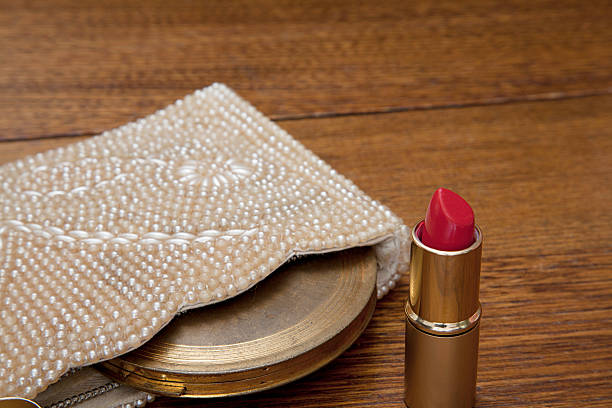 Open red tube of lipstick sitting by 1020s purse with antique powder compact on old oak dressing table. Closeup