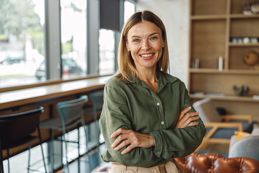 Stylish middle-aged businesswoman standing with crossed hands on coworking space background