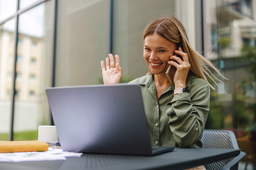 Stylish woman freelancer talking with client via video call and waving Hi while sitting outdoors