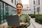Woman entrepreneur working on laptop outside on modern building background and looking camera