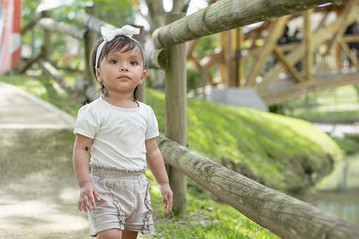 little brown-skinned latina girl, standing in the middle of the path looking curiously and very serious towards the front.