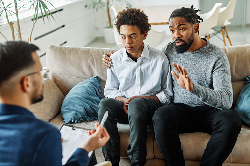 Father with his teenage son at meeting with social worker, psychologist discussing mental health family sitting on sofa in psychotherapist office