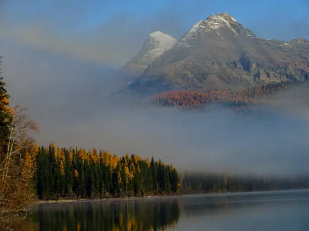 Snow capped mountain peaks peer out of the rising mysterious mist on Whiteswan Lake, BC, in Fall with trees turning orange
