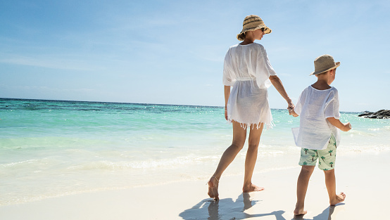 Back view of happy mother and son walking, relaxing on the tropical sandy beach, Enjoying life. Summer day. Vacation. Travel.