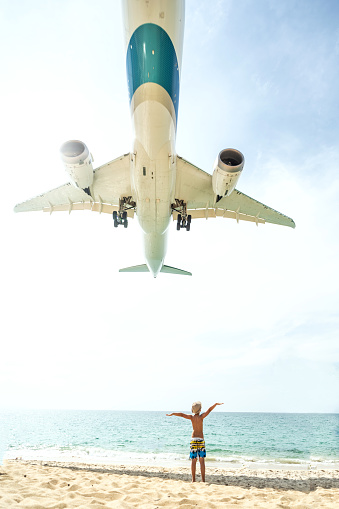 Photo of airplane flies extremely low over the sandy beach and one tourist kid. Travel and tourism concept. Thailand.