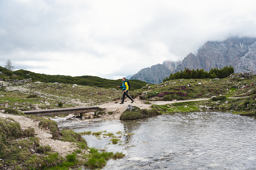 Caucasian man, crossing the small wooden bridge above the stream, while admiring view of Dolomites, during his hike