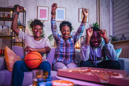 Group of young cheerful friends cheering for their sports team while watching a basketball game at home