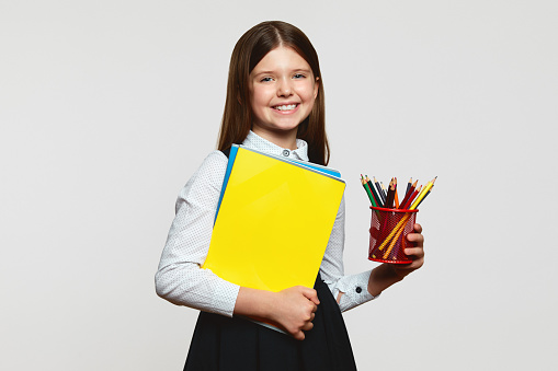 Little nerdy girl in school uniform holding notebook and red cup with multicolored pencils while standing against white background. Back to school