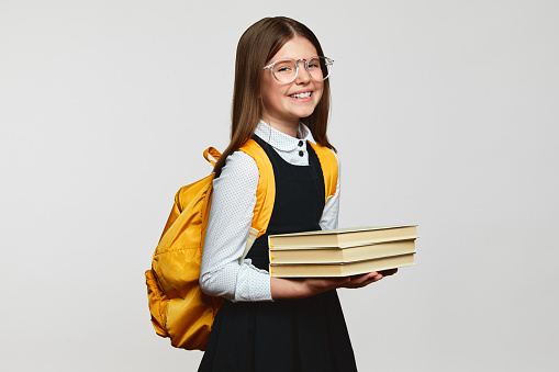 Positive smart school girl wearing eyeglasses and yellow backpack holding books and smiling at camera against white background. Back to school concept