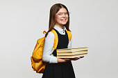 Positive schoolgirl with eyeglasses and yellow backpack holding bunch of books in hands and smiling