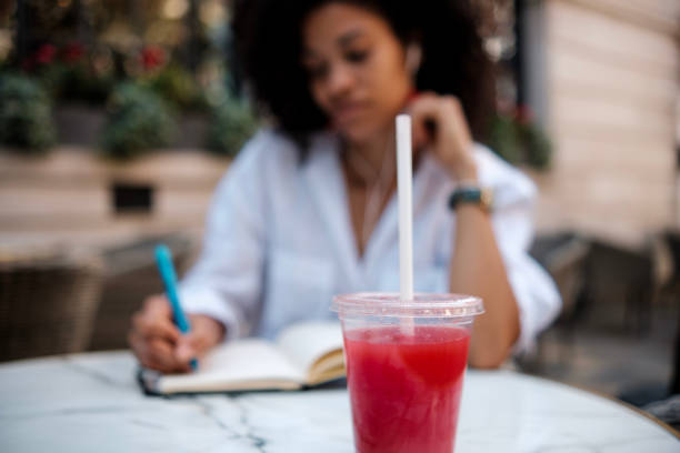 Close-up of juice cup with blurred woman writing on notebook. Tasting and drinking sweet beverages in hot summer. Alcohol free, healthy drinks concept.
