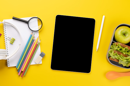 Tablet with blank screen, school supplies, stationery, and lunch box on yellow background. Education and nutrition. Flat lay with blank space for your text or app