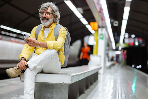 Shot of a modern senior man in a yellow shirt with a backpack waiting for a train at the metro station. He is holding a smart phone