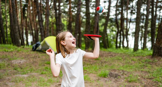 Little girl playing in the camping in the wood with ball and laughing at the nature with green tent on background. Beautiful smiling kid having activities in the forest with sun light