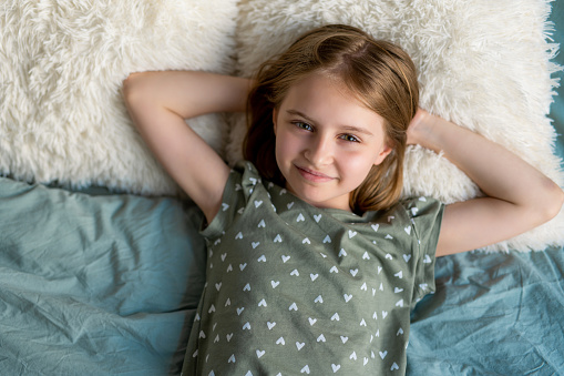 Little beautiful kid girl with blond hair lying in the bed and looking at the camera in bedroom with white furry pillows. Schoolgirl resting at home