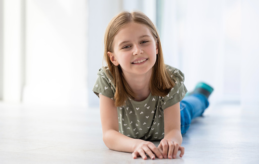 Portrait of preteen beautiful girl lying on the floor in light room and smiling. Female kid schoolgirl looking at the camera indoors