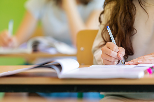 Teenage girl sitting at the desk in the classroom during lesson, writing in notebook. Close up of hand, unrecognizable person.