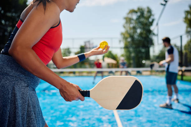 Black female pickleball player serving the ball during the match on outdoor court. Unrecognizable African American woman serving the ball while playing mixed doubles in pickleball. pickleball equipment stock pictures, royalty-free photos & images