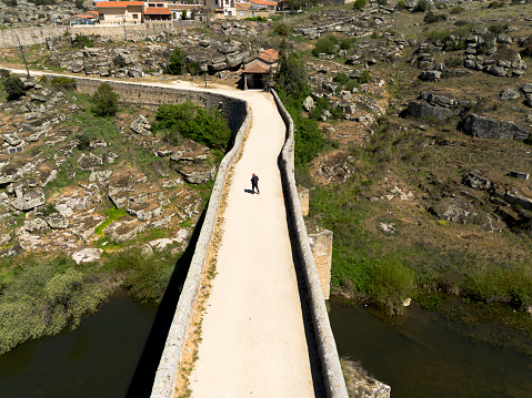 A middle-aged man, on a medieval bridge, with a black backpack, is in the middle of the sand bridge, where at the end there is a small church, old bridge (Ledesma) - Salamanca.