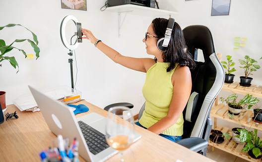 Smiling young woman wearing headphones adjusting her smart phone while doing a vlog on a laptop at a desk in her home office