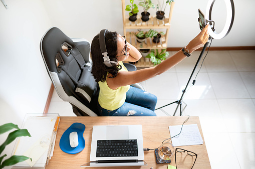 High angle view of a young woman wearing headphones adjusting a smart phone while setting up for a vlog at a desk