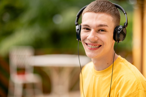 Portrait of a young man with braces smiling while listening to music on headphones outside on a patio at home