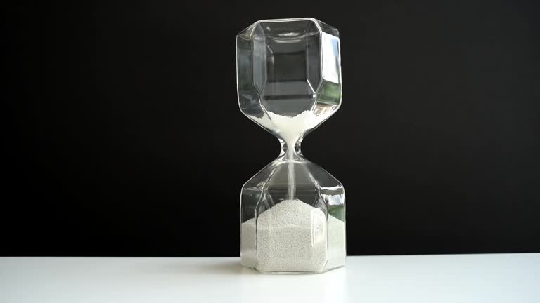 Hourglass on a white table and black background. Faceted modern hourglass with light gray sand. The last grains of sand are pouring out