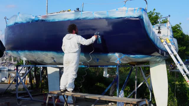 A worker in a body protective suit paints the hull of a sailing yacht with spray.