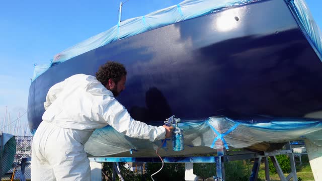 A worker in a body protective suit paints the hull of a sailing yacht with spray.