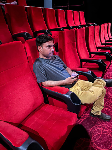 Stock photo showing close-up view of man sitting in mid row seat of cinema wearing with on leg crossed on lap to watch a film.