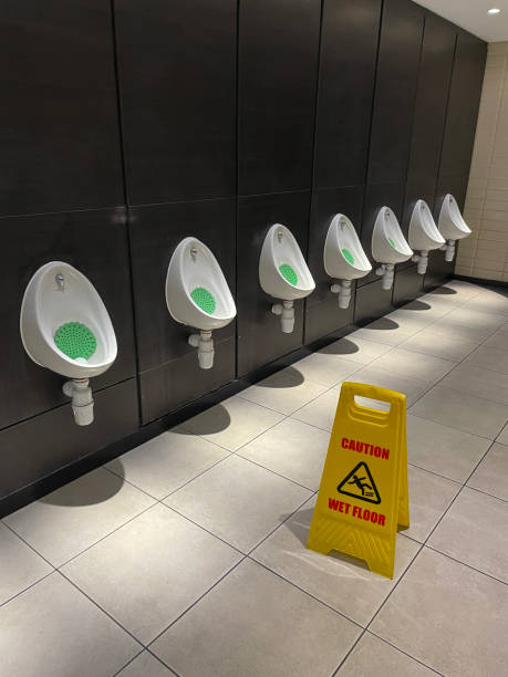 Stock photo showing close-up view of yellow and red, rigid plastic, free standing advisory A-frame sandwich board 'Caution Wet Floor' sign before row of urinals in public washroom toilets with green PVC, scented urinal screen located in restroom urinal bowls. Impregnated with a fragrant deodoriser the PVC mats are designed to keep the urinal fresher smelling. imagen del letrero independiente