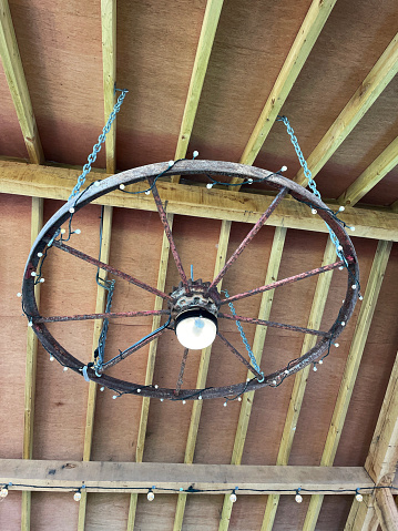 Stock photo showing an iron rimmed wagon wheel hanging from barn ceiling by chains. The wheel has been upcycled into a lighting design lighting by wrapping it in electric string light bulbs.