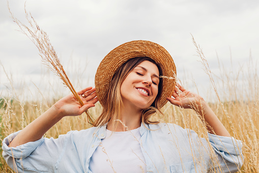 Portrait of happy young woman walking among high grasses in summer meadow wearing straw hat and linen shirt picking bundle of dry grass. Harmony and balance. Wellbeing