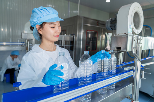 Unveil the essence of perfection in the manufacturing industry as a worker woman meticulously examines water bottles. Wearing lab coat symbolizing expertise and concentration reflecting unwavering dedication. Ensures that only the finest bottles proceed on the conveyor belt.
