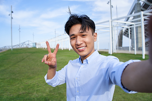 Selfie portrait looking at camera of Asian teenage boy making peace sign. Smiling attractive young man standing outdoors with grass in background. Happy people positive emotions and social networks.